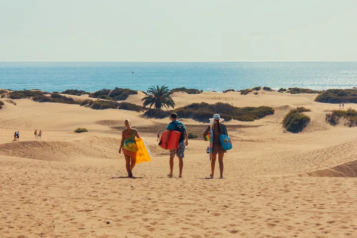 Tre personer med badeutstyr går over sanddynene i Maspalomas
